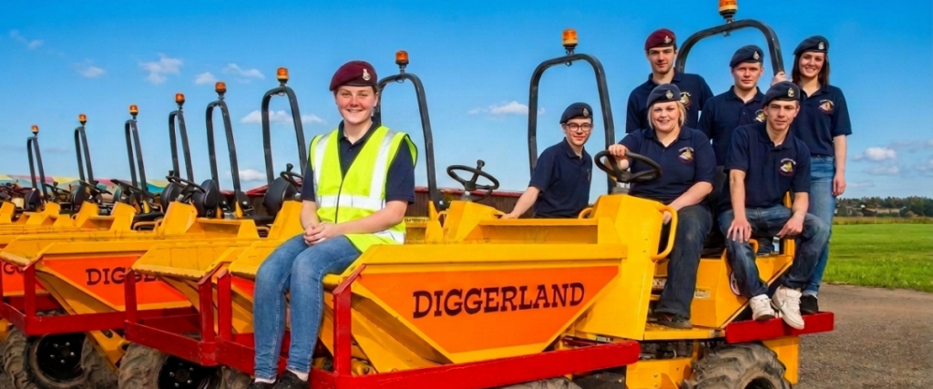 Diggerland staff members wearing navy blue polo shirts and cadet berets posing on yellow dumpers for National Cadets Week 2026, with the front staff member wearing a high-visibility safety vest.